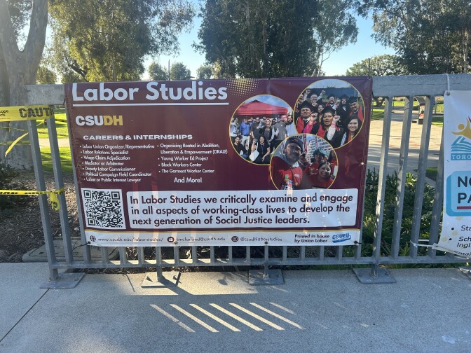 A burgundy sign titled "Labor Studies" is affixed to a gate on a college campus. It features photos of students in black caps and gowns, along with a description of the types of careers and internships available to students who pursue this field.