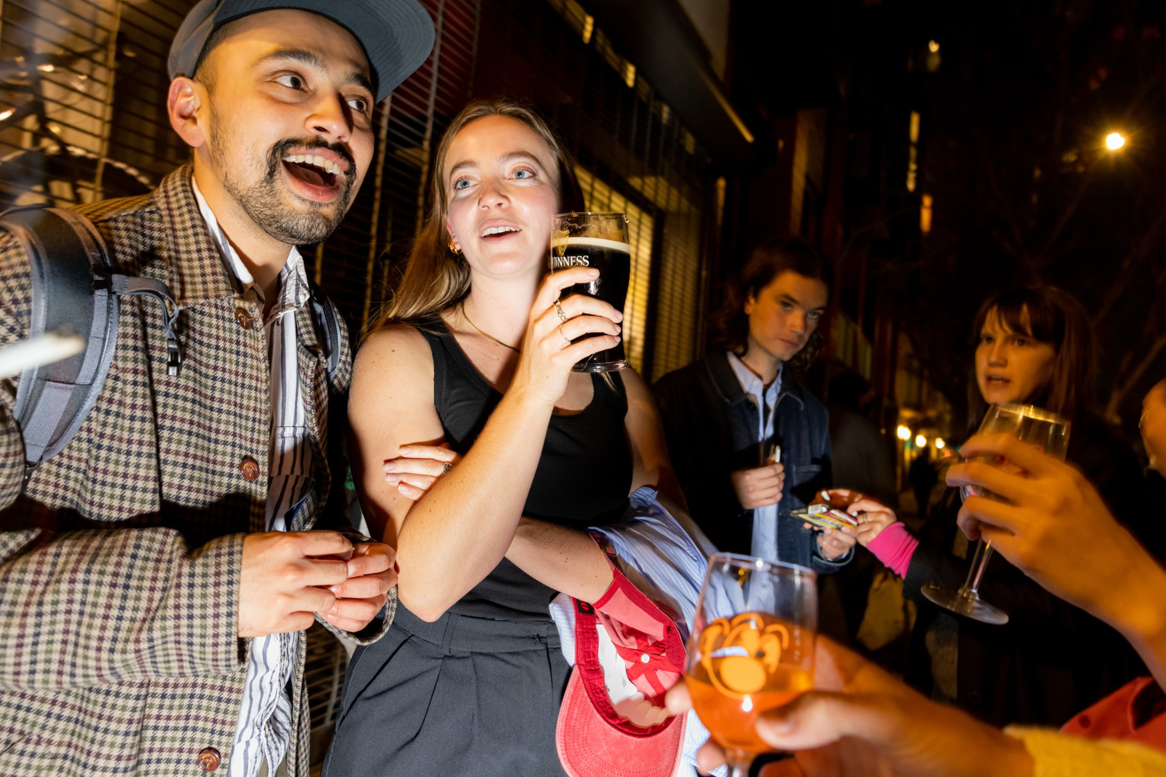 A group of young people stand outside at night, holding drinks and chatting, with one woman holding a glass of dark beer and others holding wine glasses.