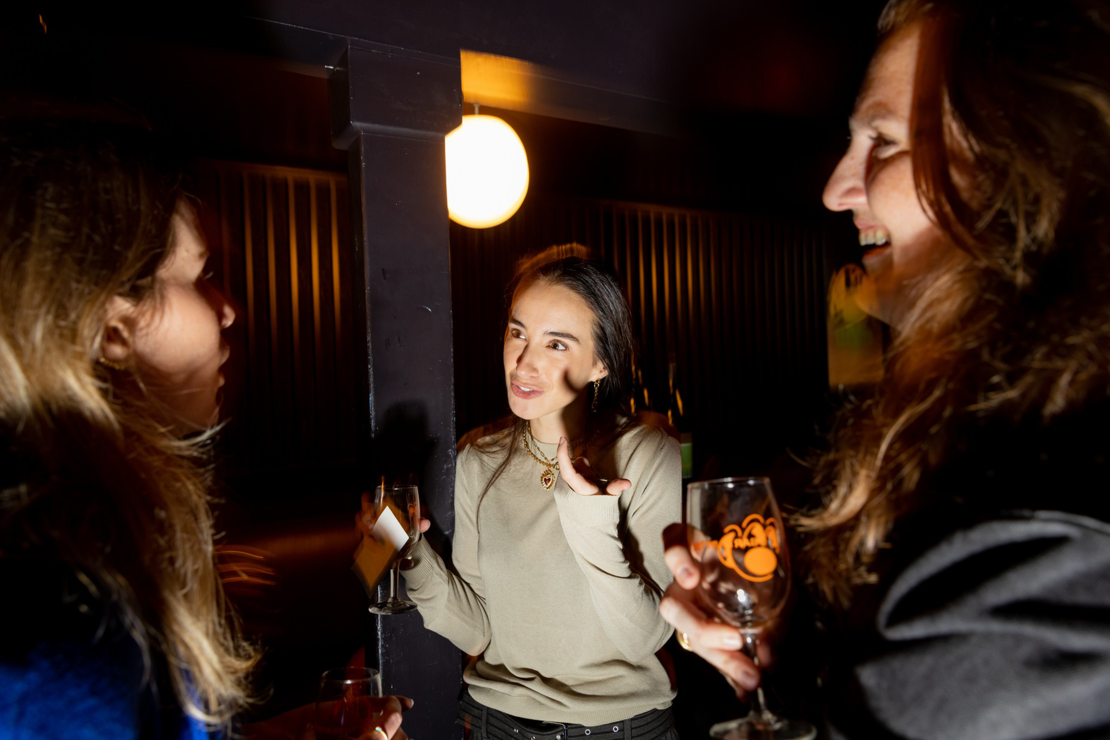 Three women are engaged in conversation in a dimly lit room, holding drinks and smiling, with a round light glowing in the background.
