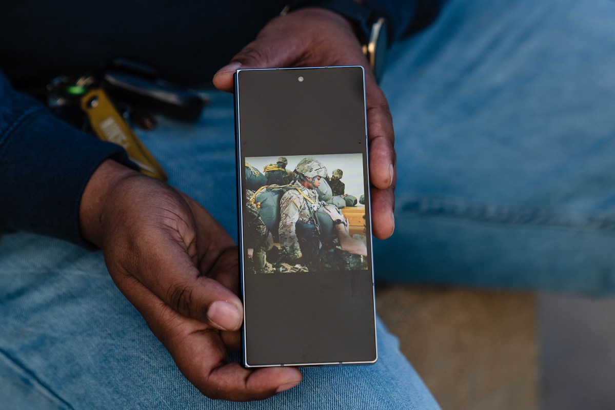 A person holds a smartphone in both hands while seated, showing a photo on the screen of several soldiers in military gear clustered together outdoors, with the person’s jeans and a set of keys visible in the background.