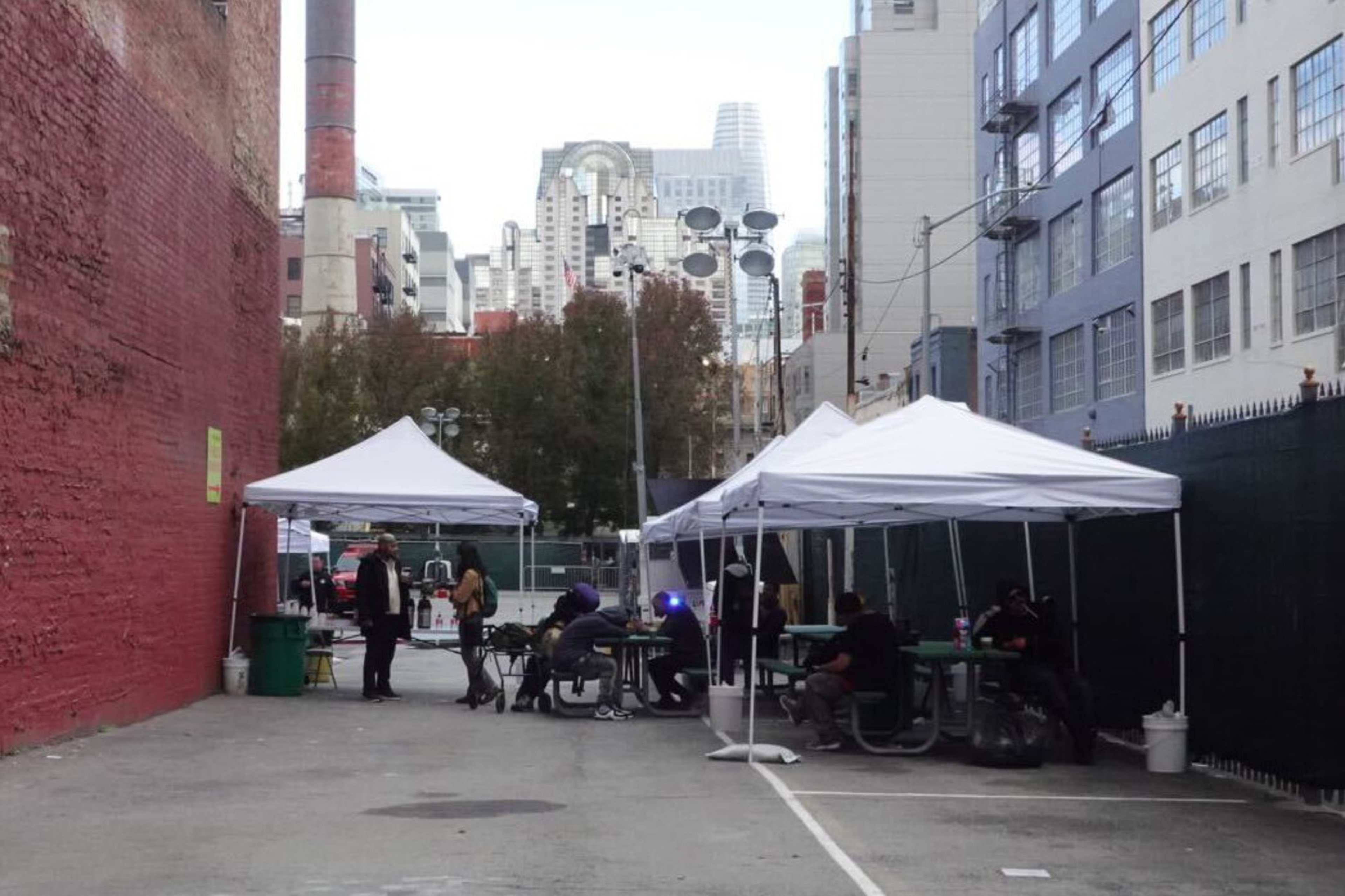 An alley with white tents covering tables, where a few people sit and stand. Tall buildings and trees are visible in the background.