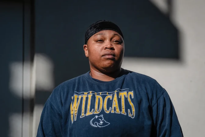 A woman with dark skin tone, wearing a dark blue long-sleeve shirt with text that reads "West L.A. Wildcats." looks out of frame while standing in front of a building that is partially covered in shade.