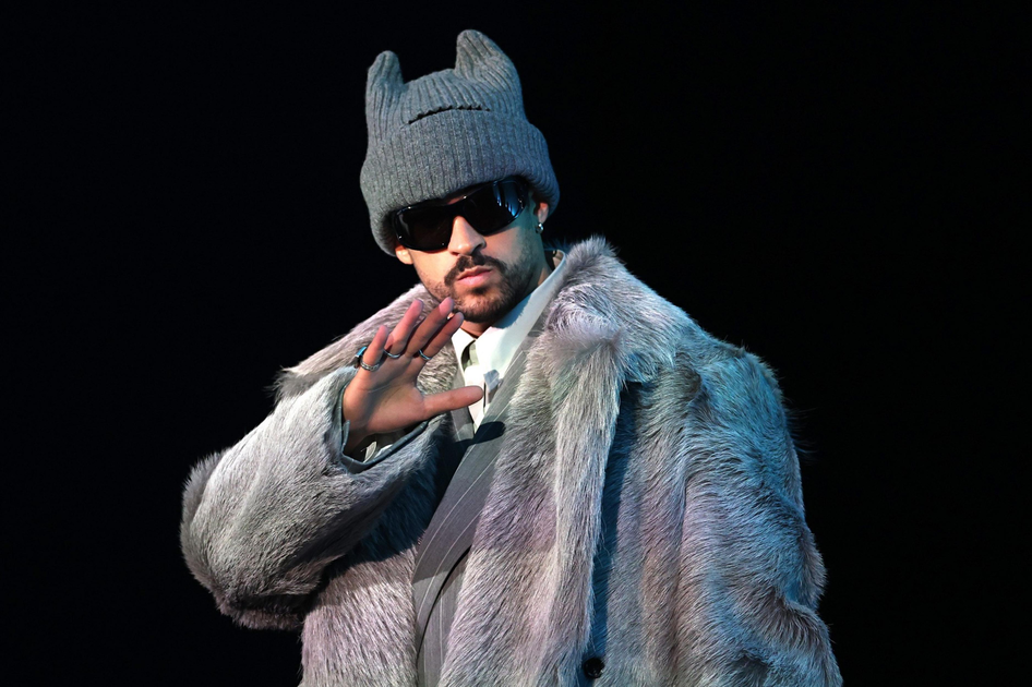 A person wearing a large gray fur coat, a gray knit hat with cat ears, dark sunglasses, and rings raises one hand near their face against a black background.