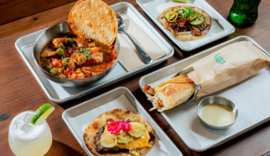 A meal spread includes a bowl of stew with a large tortilla chip, two tacos with various toppings, a wrapped sandwich, a creamy dipping sauce, a green bottle, and a lime drink.