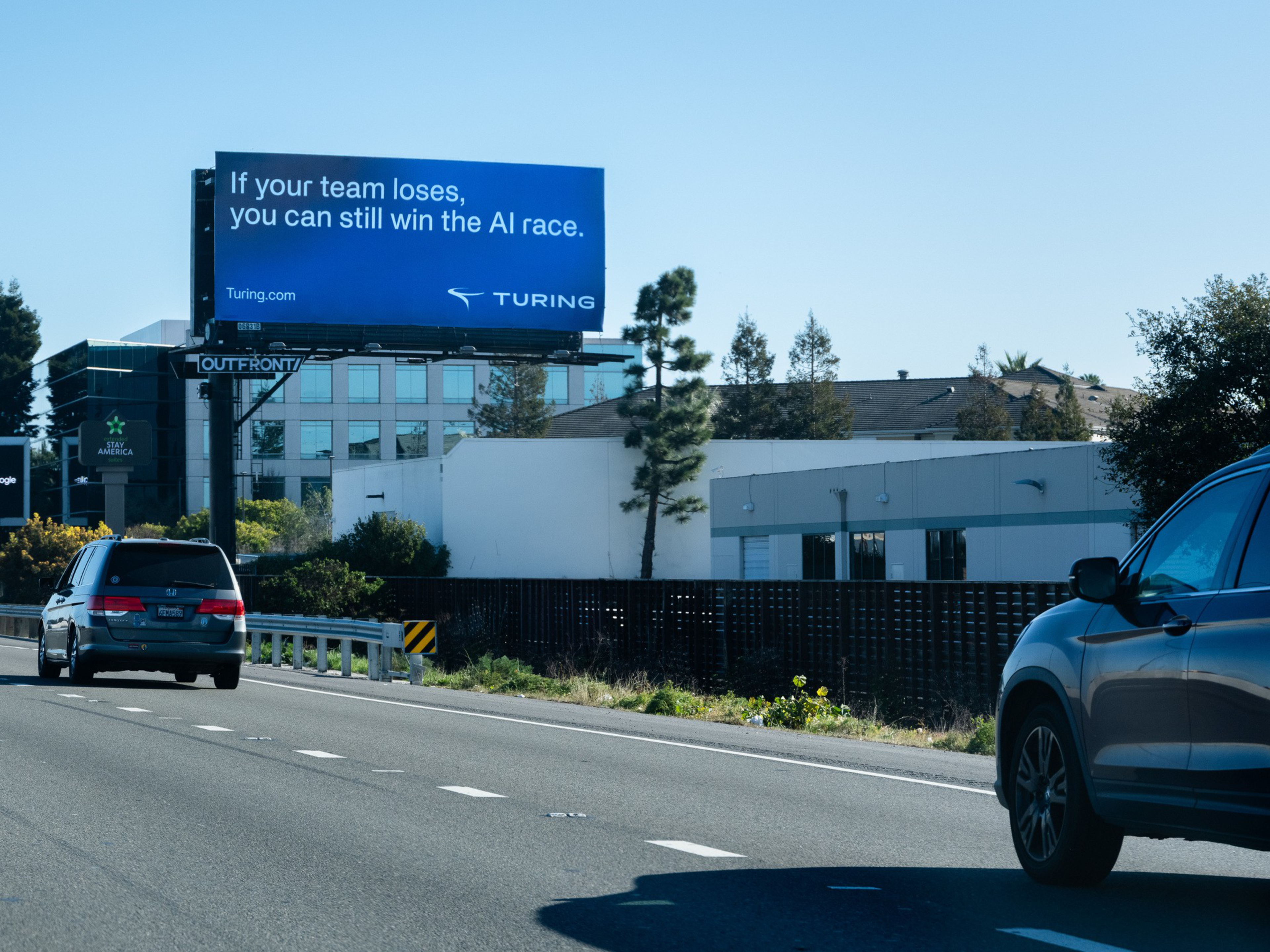 A blue billboard by a highway reads, “If your team loses, you can still win the AI race,” with the logo and URL for Turing.com. Several cars are on the road.