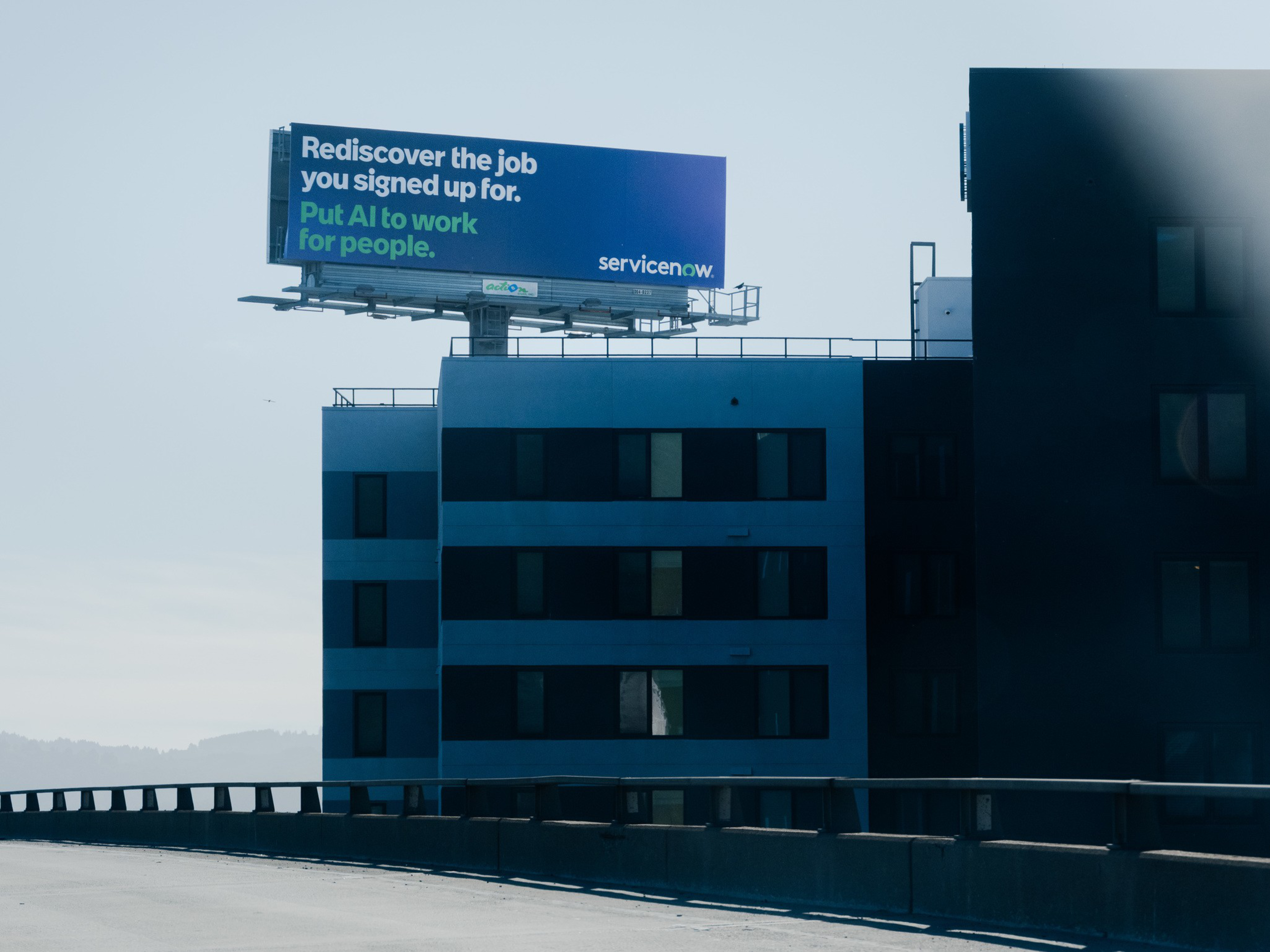 A billboard atop a building reads, “Rediscover the job you signed up for. Put AI to work for people. servicenow.” The sky is clear.
