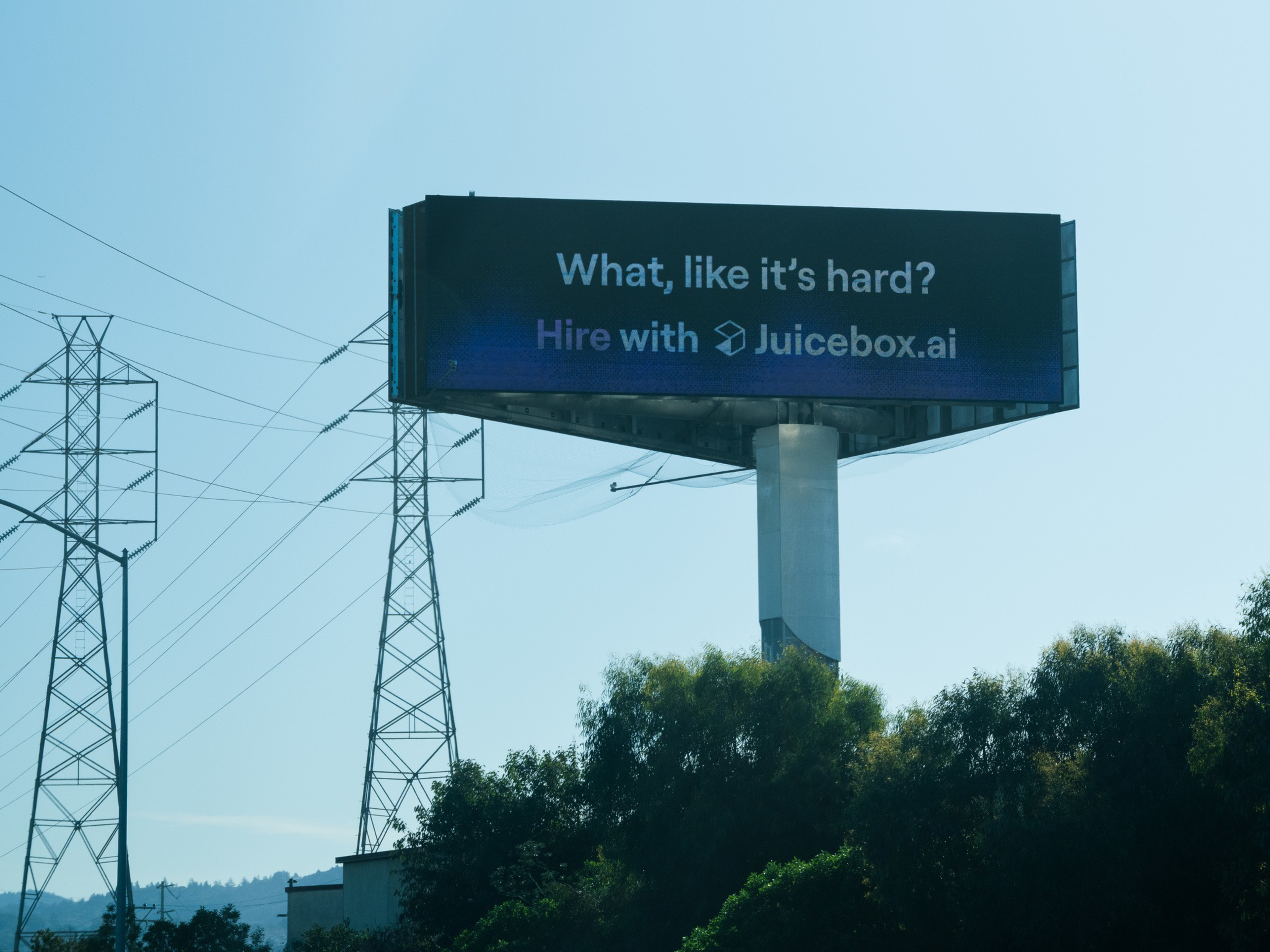 Billboard text reads: “What, like it’s hard? Hire with Juicebox.ai” against a clear sky with power lines and trees below.