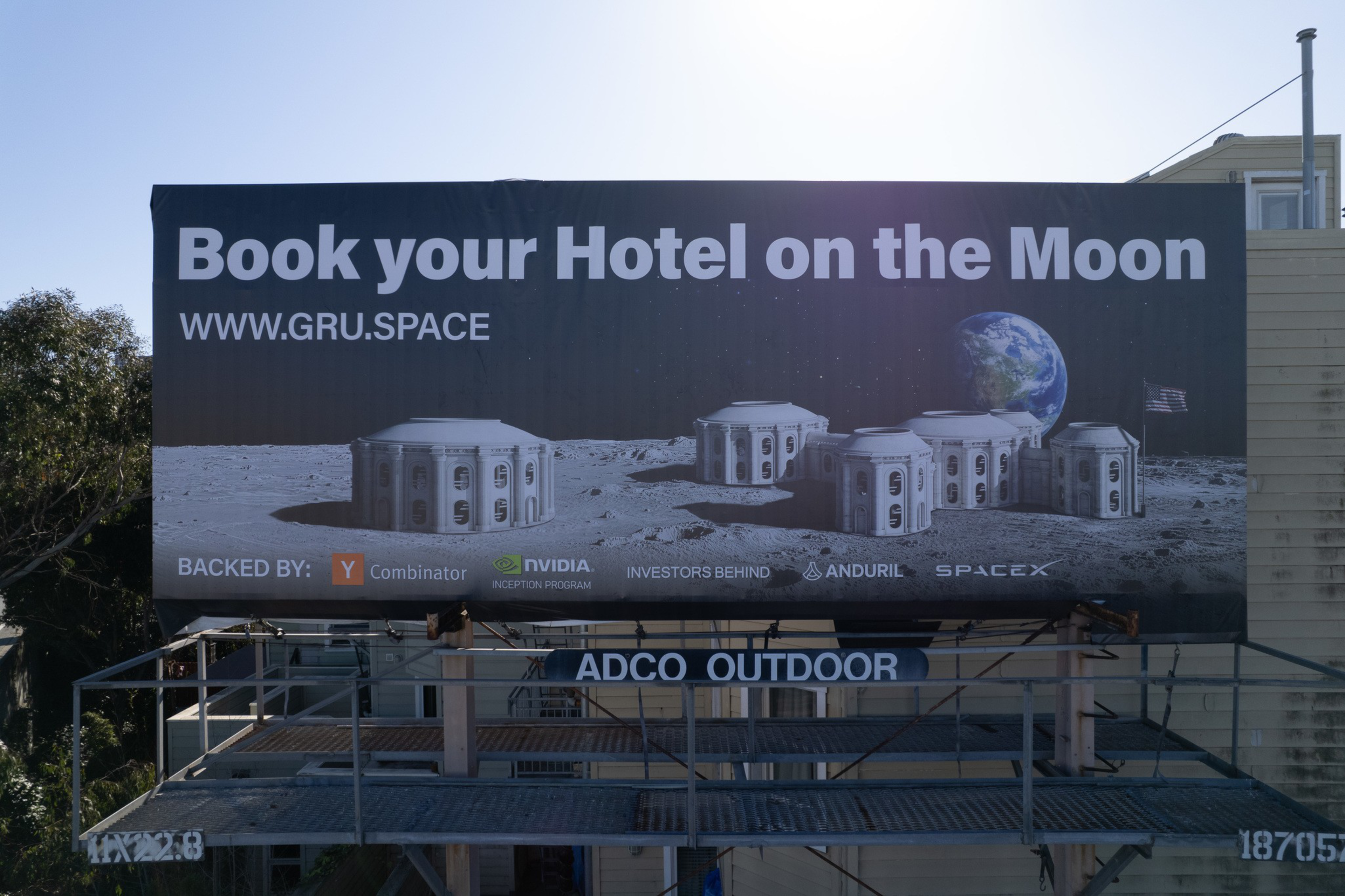 A billboard advertises booking a hotel on the Moon at www.gru.space, showing futuristic lunar buildings with Earth and a flag in the background.