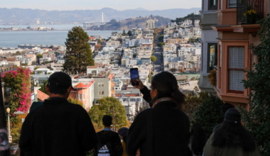 People stand on a steep street overlooking a dense cityscape with water and hills in the background under a clear sky.