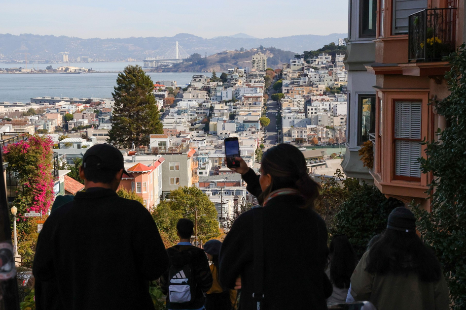 People stand on a steep street overlooking a dense cityscape with water and hills in the background under a clear sky.
