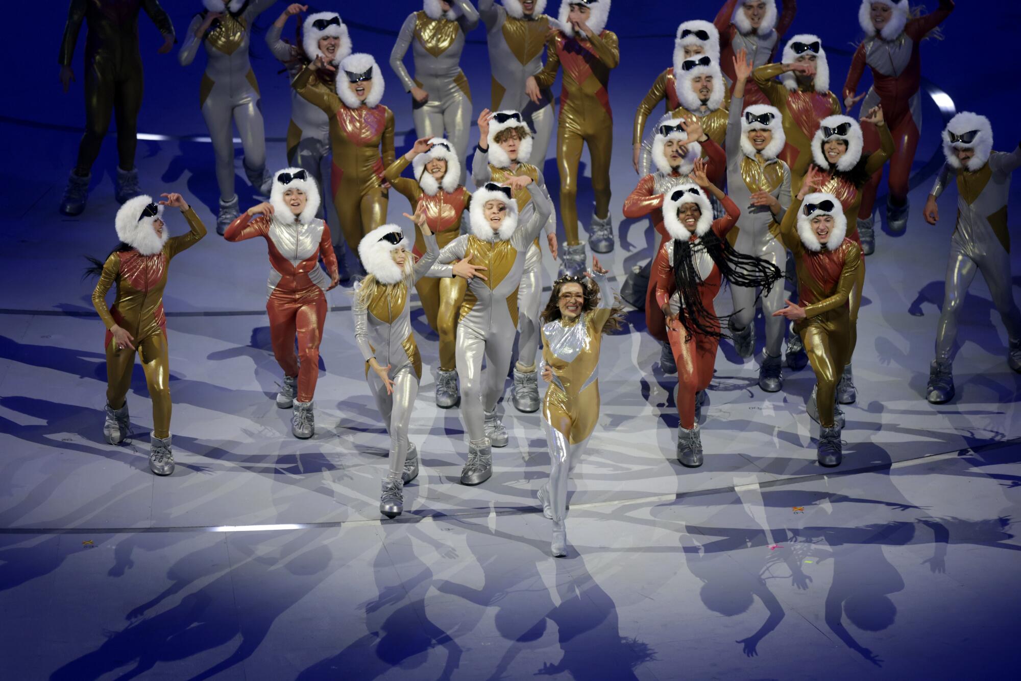 A woman in a silver and gold leotard surrounded by dancers on a stage.