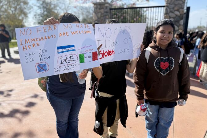 Three students stand in a group. Only one has her face visible; she has medium skin tone and wears a brown sweatshirt with a heart on it. The two students next to her hold up signs in front of their faces, with phrases like "ICE out now" on them.