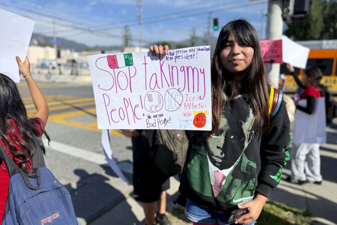 A girl with long dark hair and medium light skin tone holds up a sign that says "Stop taking my people!" while students hold up signs around her.