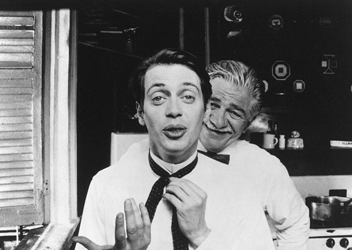 A man fixes his tie while another speaks over his shoulder.