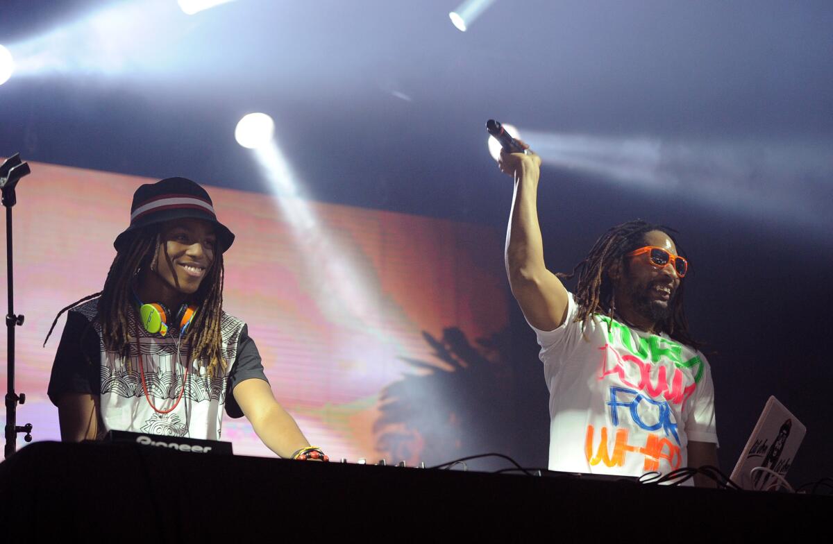 A man in a hat performing at a DJ booth while another man holds up a microphone next to him