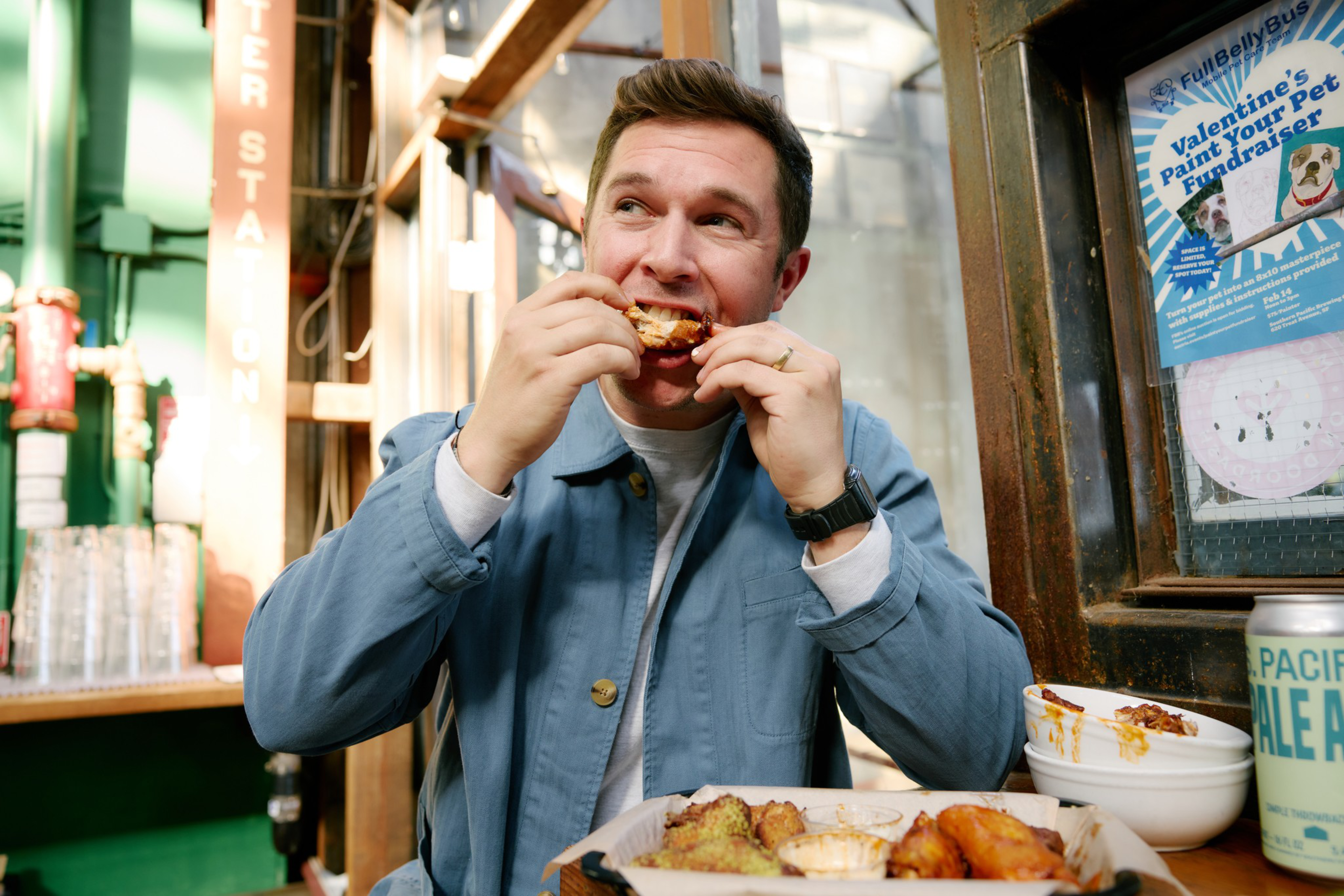 A man wearing a blue jacket eats chicken wings at a table with food, dipping sauces, a drink can, and a fundraiser poster on the wall behind him.