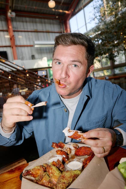 A meal spread includes a bowl of stew with a large tortilla chip, two tacos with various toppings, a wrapped sandwich, a creamy dipping sauce, a green bottle, and a lime drink.