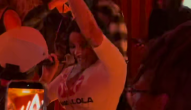 A woman sings energetically into a microphone, raising her arm, while people around her, including someone recording on a phone, watch in dim red lighting.