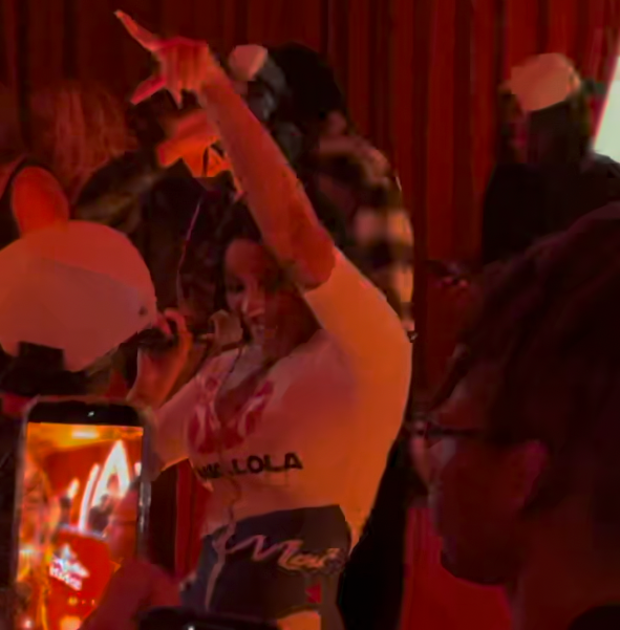 A woman sings energetically into a microphone, raising her arm, while people around her, including someone recording on a phone, watch in dim red lighting.