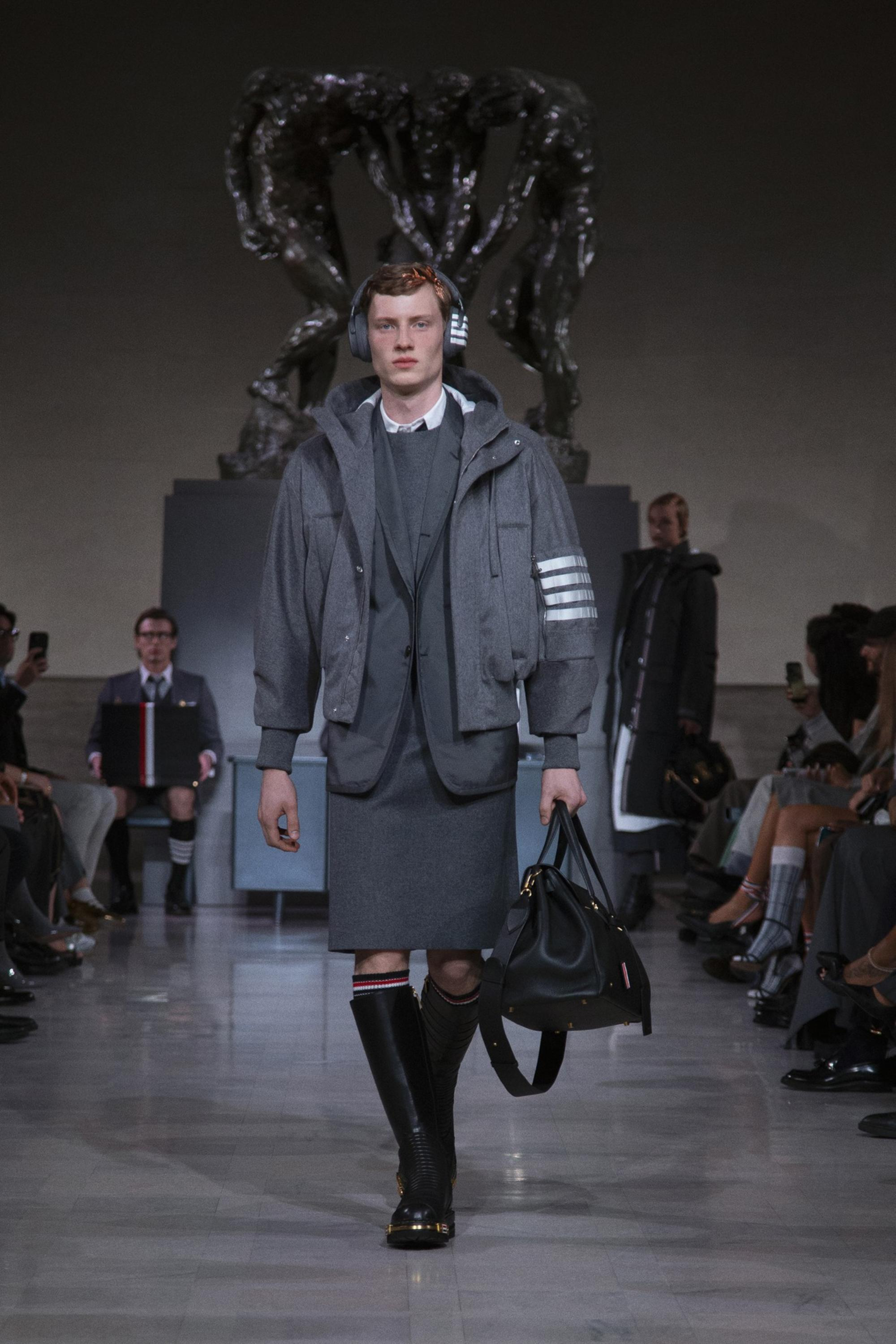 A male model walks a runway wearing a gray layered outfit with a skirt, a jacket, knee-high boots, headphones, and carrying a large black bag.