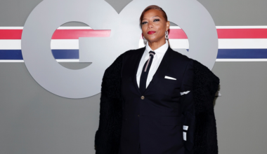 A woman in a black suit with a white shirt, striped tie, and black fur coat poses confidently against a gray background with a large "GQ" logo.
