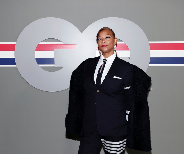 A woman in a black suit with a white shirt, striped tie, and black fur coat poses confidently against a gray background with a large "GQ" logo.