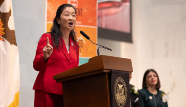 A woman in a red suit speaks passionately at a podium with a seal. A flag is partially visible behind her, and another person is seated in the background.