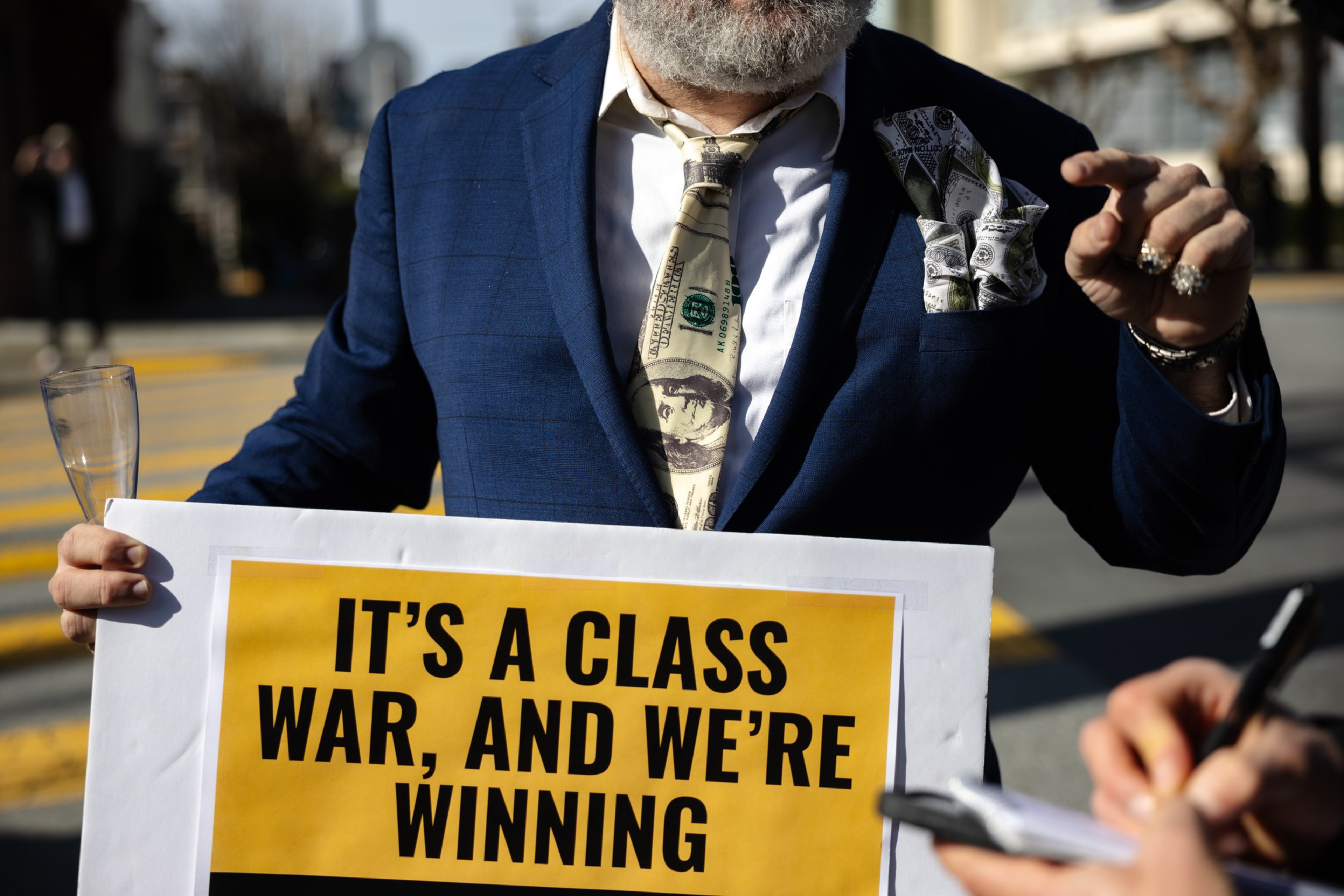 A person in a blue suit holds a sign that reads “IT’S A CLASS WAR, AND WE’RE WINNING,” while wearing a money-print tie and pocket square.