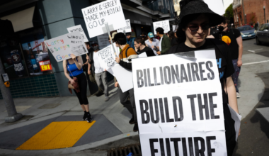 People march in a street protest holding signs with messages like "BILLIONAIRES BUILD THE FUTURE" and "LARRY & SERGEY MADE MY 401(K) GO UP."