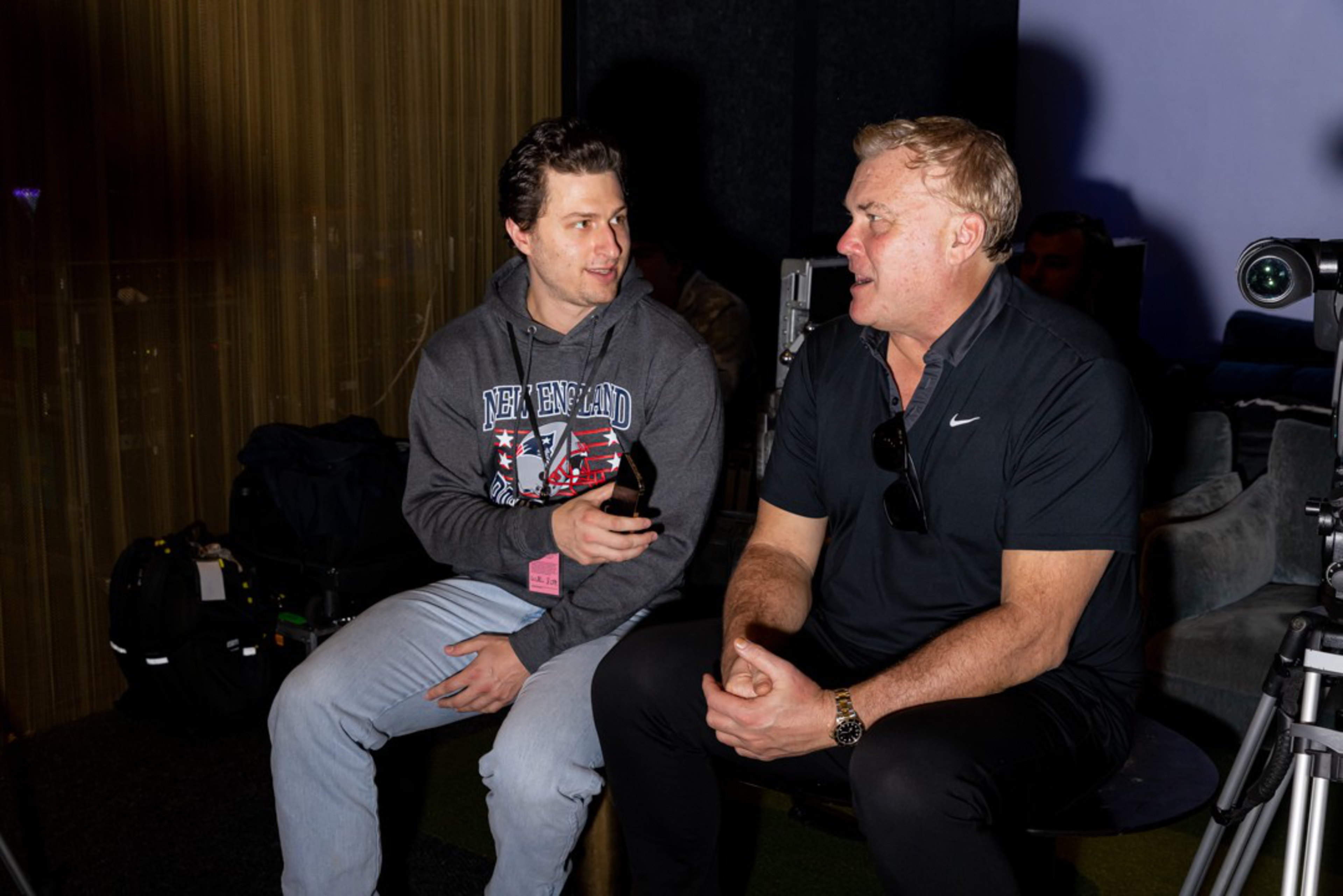 Two men are sitting and talking; one wears a gray New England hoodie and light pants, while the other wears a black Nike polo and dark pants.