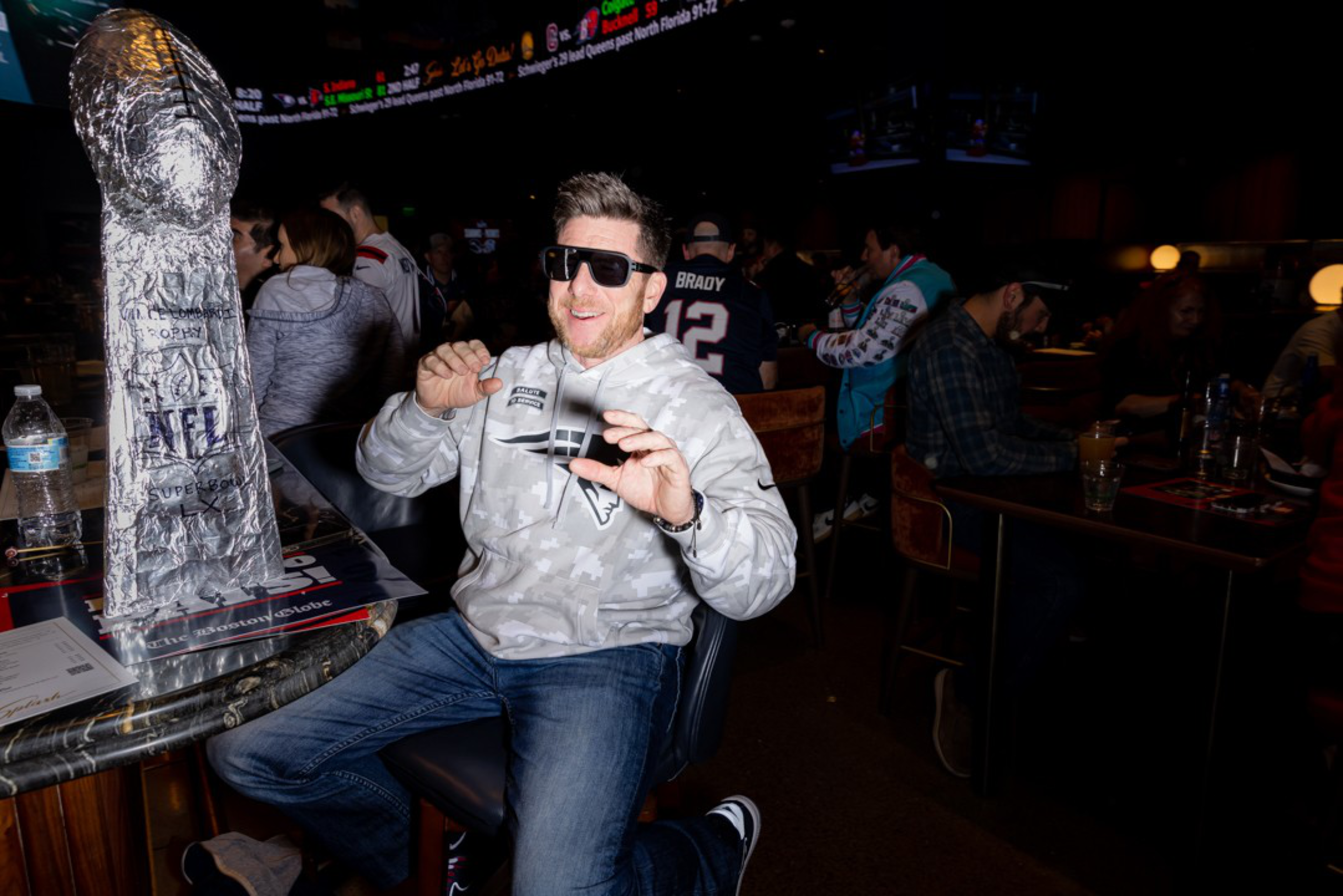 A man wearing sunglasses and a gray Patriots hoodie poses excitedly next to a large foil-wrapped object resembling a football trophy on a table.