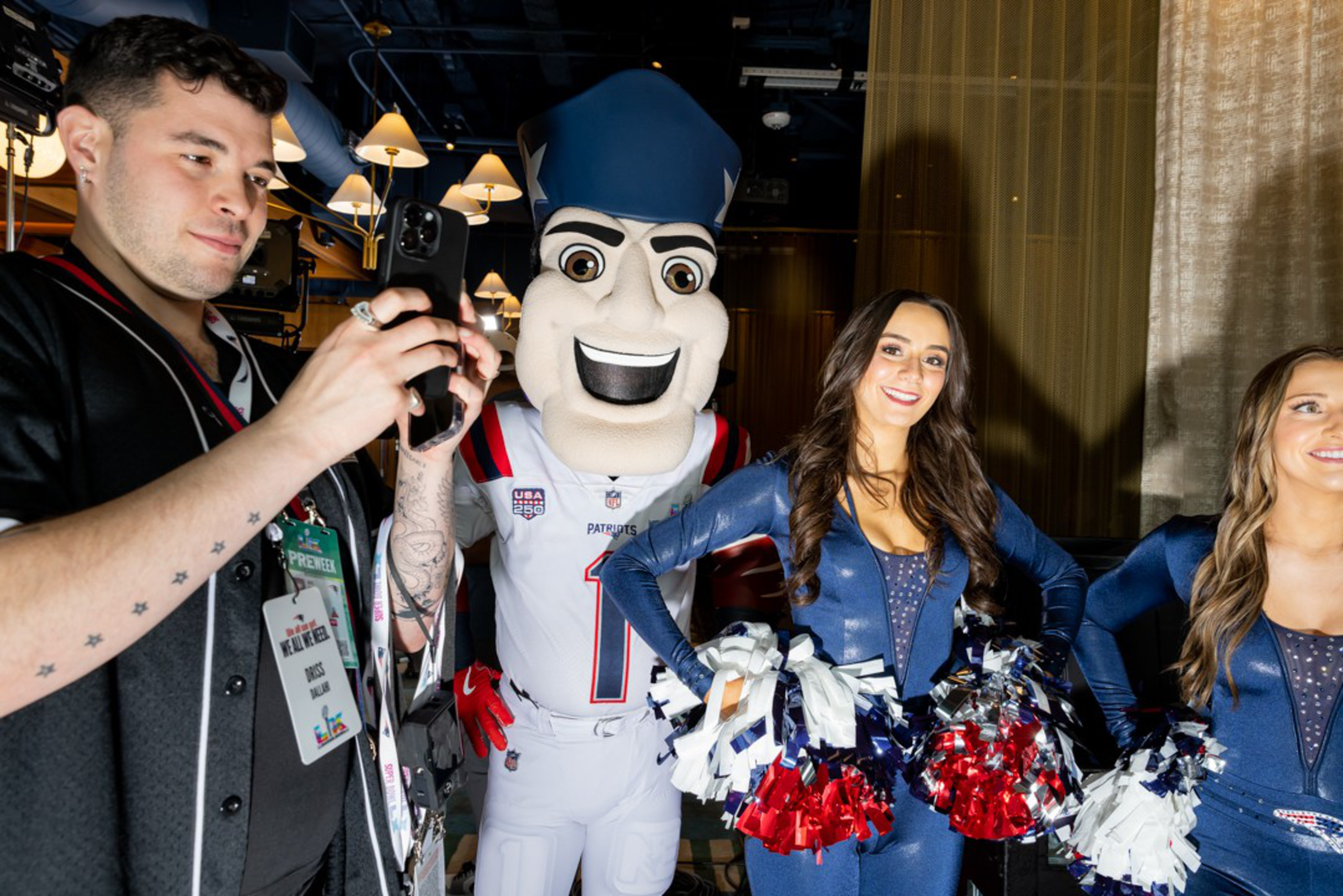 A man takes a photo of two cheerleaders in blue outfits holding red, white, and blue pom-poms, with a Patriots mascot standing behind them.