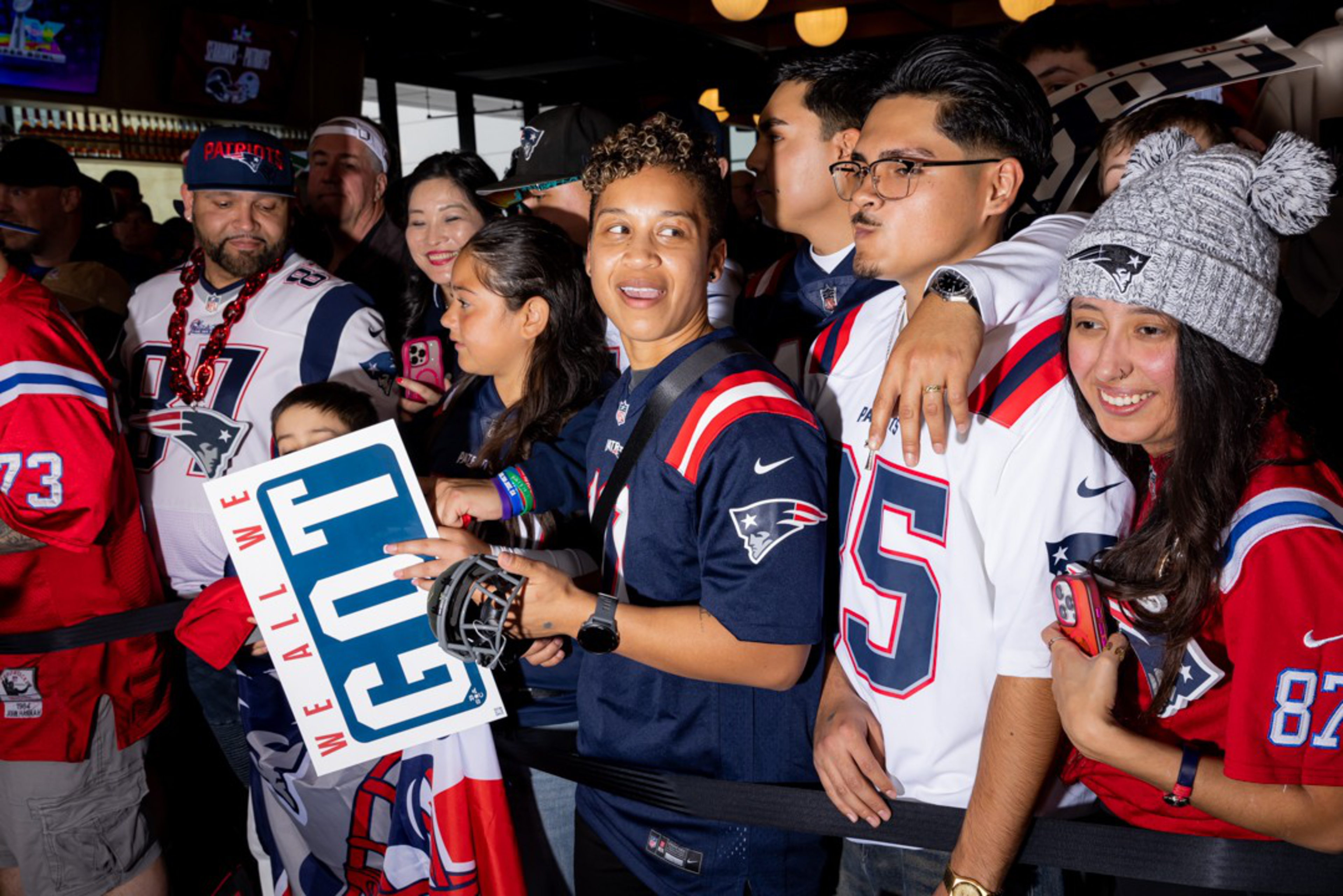 A diverse crowd of New England Patriots fans wearing team jerseys and gear, some holding signs and phones, cheer indoors at a crowded event.