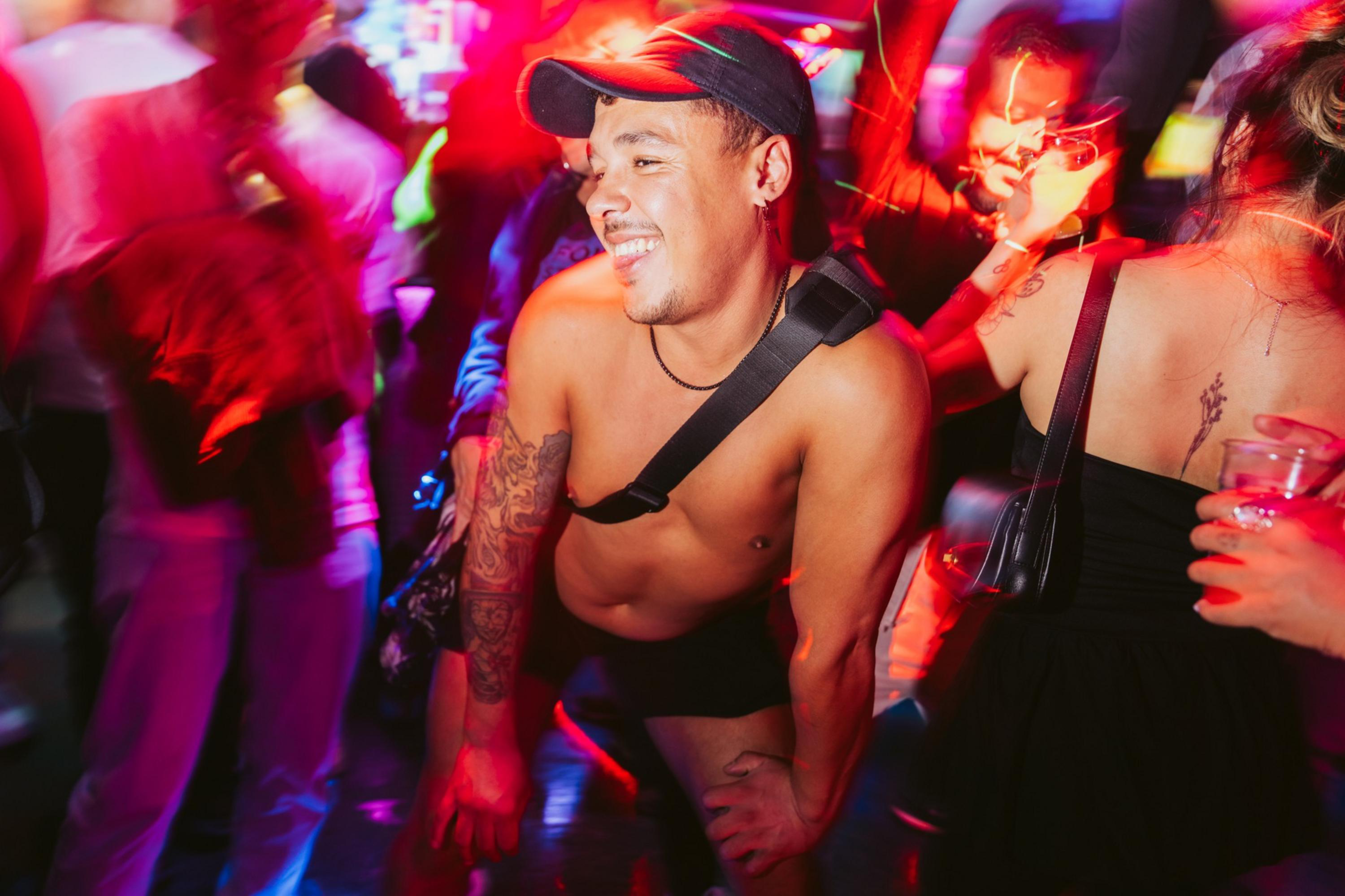 A shirtless man with tattoos and a cap smiles and leans forward while dancing in a crowded, colorful nightclub with red lighting.