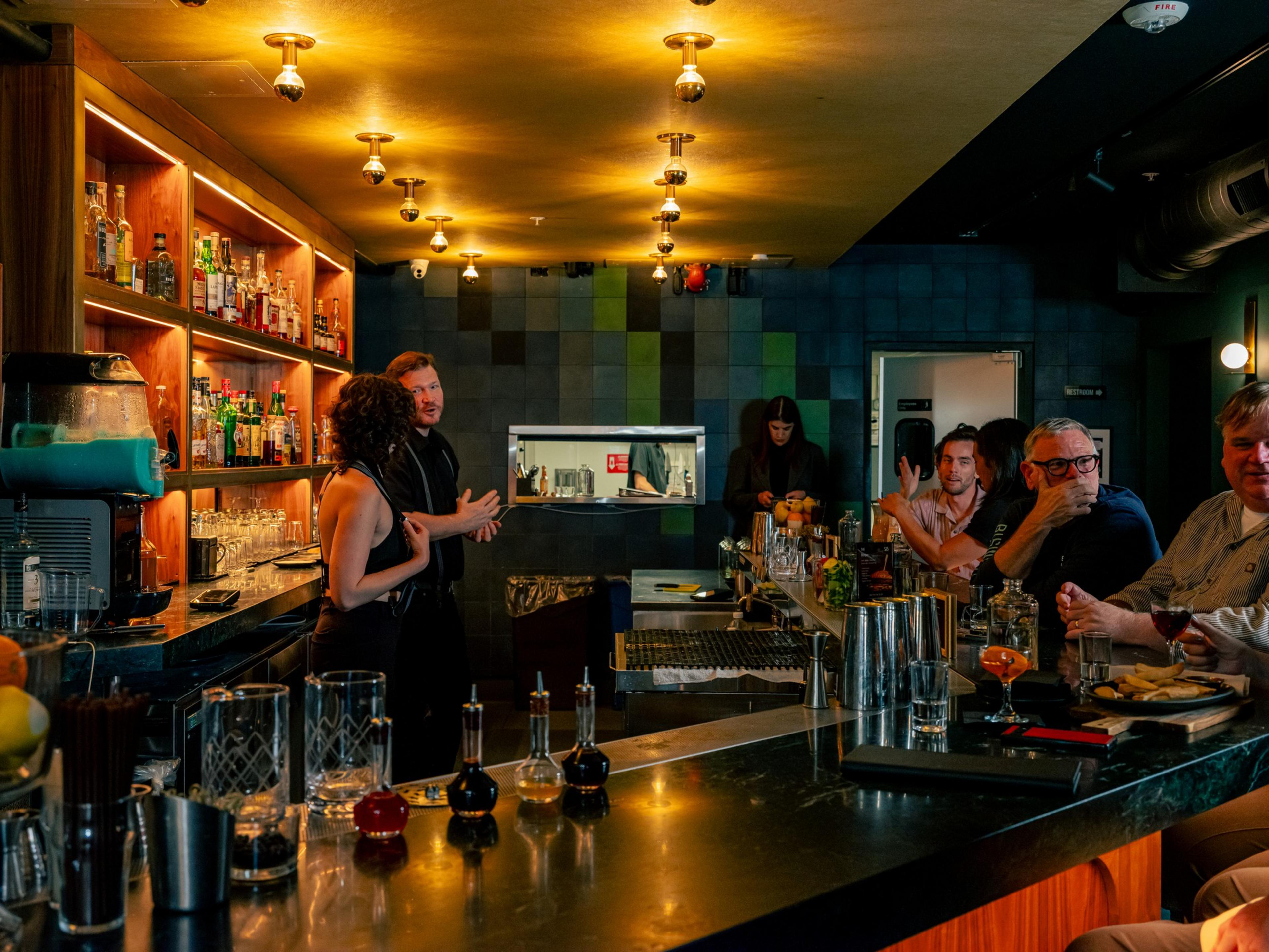 A bar with warm lighting shows a bartender and a woman talking, while four patrons sit at the counter with drinks and snacks in a cozy, modern setting.