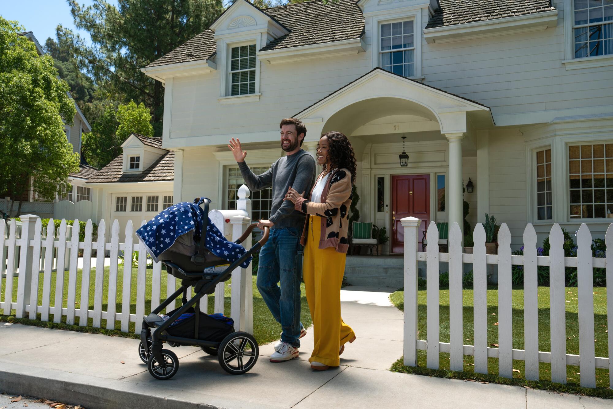 A man and a woman pushing a baby stroller outside a home with a white picket fence.