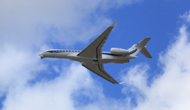 A white private jet with blue stripes flies against a partly cloudy blue sky, viewed from below and slightly behind.