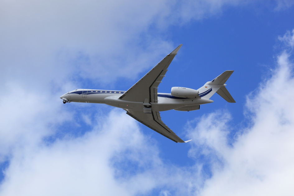 A white private jet with blue stripes flies against a partly cloudy blue sky, viewed from below and slightly behind.