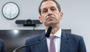 A man in a suit stands in front of a microphone, with a wall clock and a screen behind him. His expression is serious.