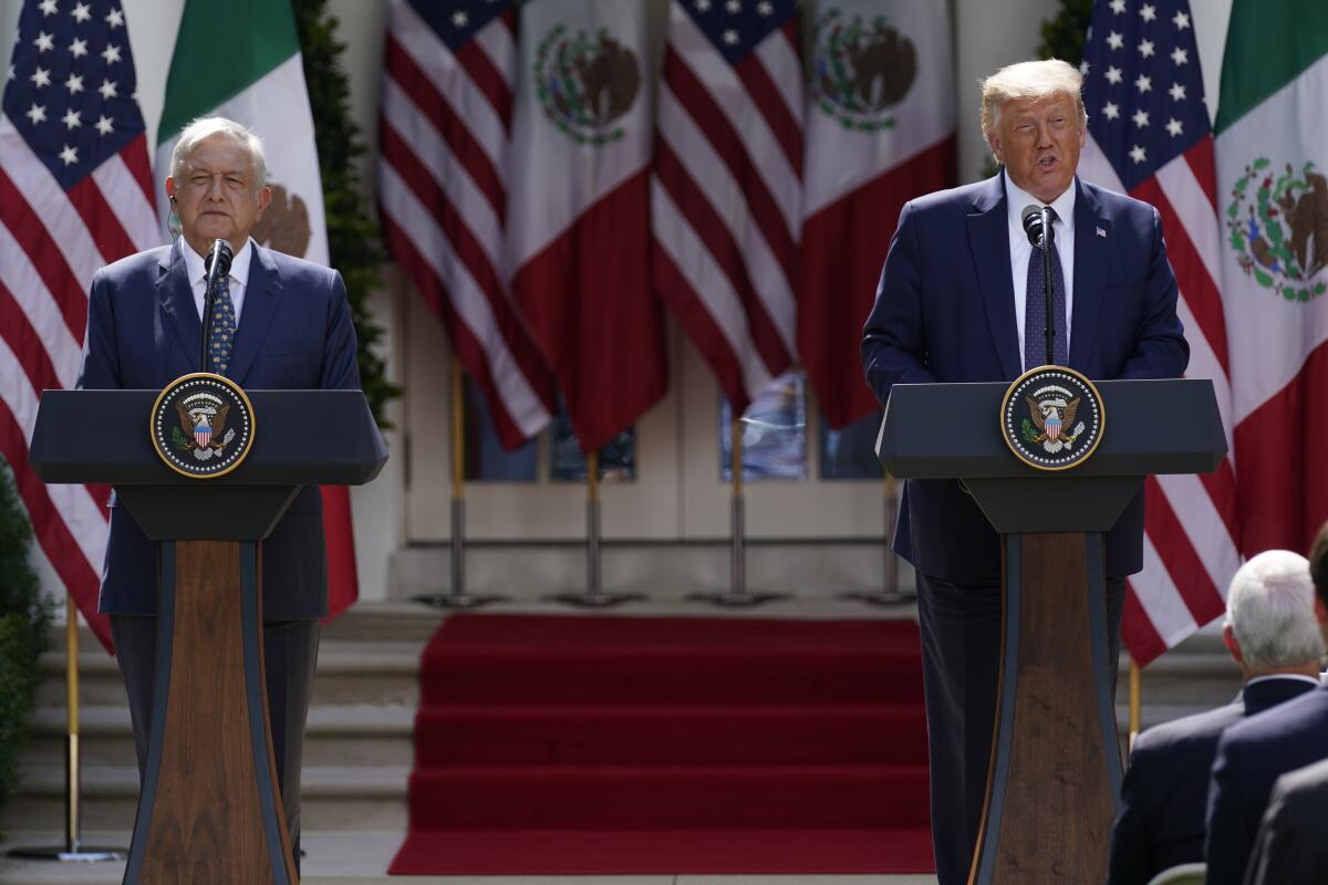 Two men in dark suits and ties standing at lecterns, with an array of flags behind them