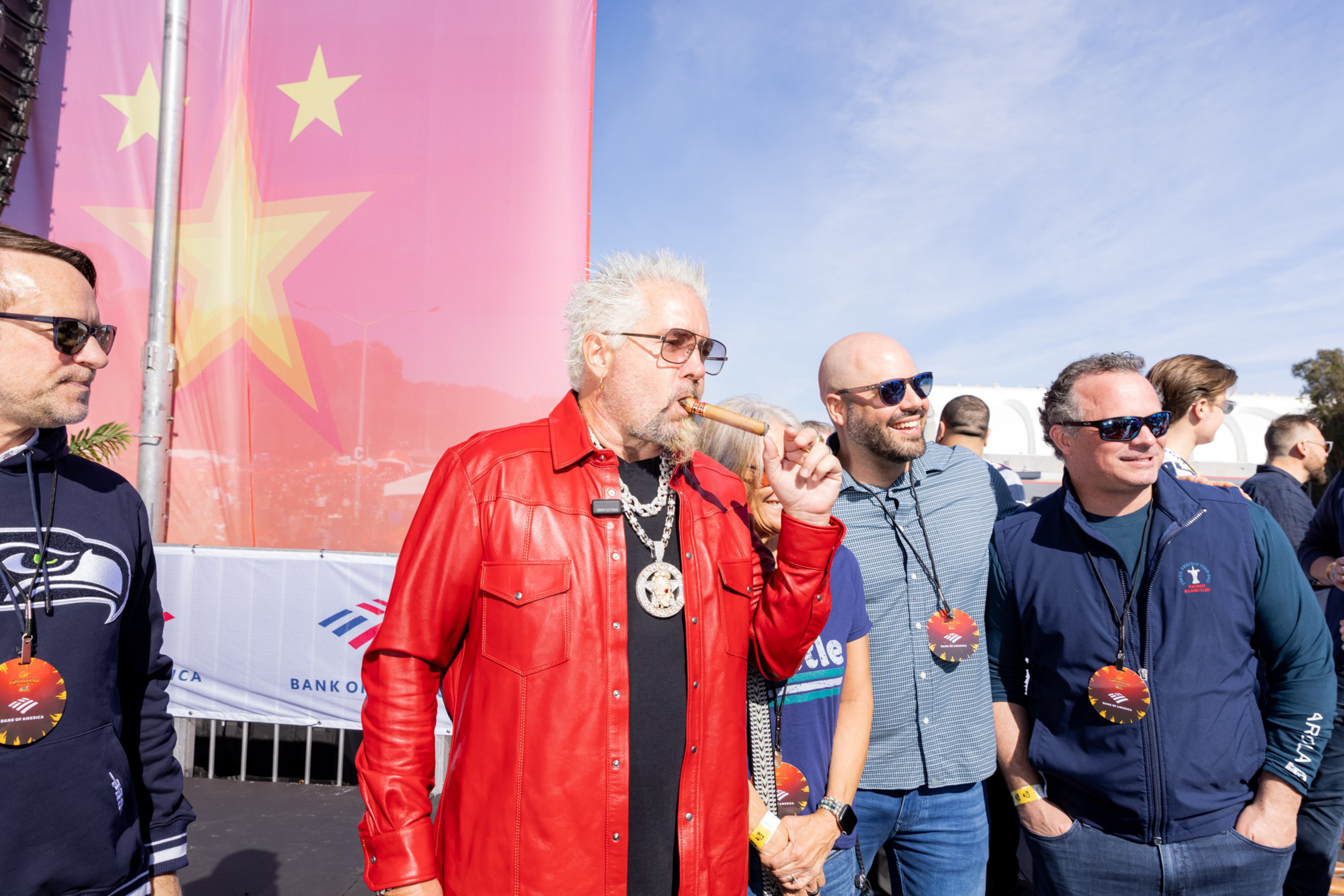 A man with white spiked hair wearing a bright red leather jacket smokes a cigar while standing with a group of casually dressed smiling men outdoors.
