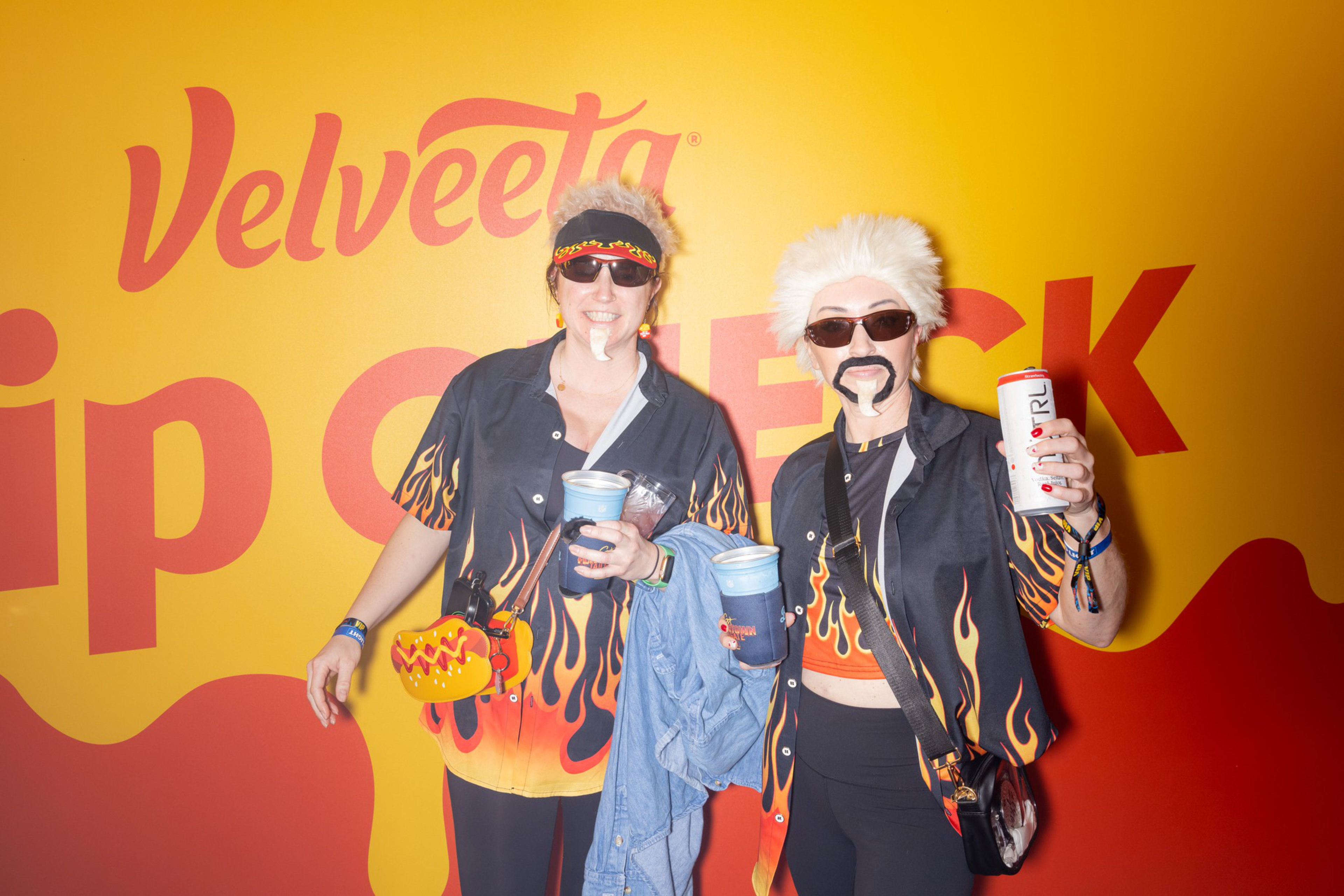 Two people dressed in matching flame-patterned shirts, sunglasses, and fake facial hair pose holding drinks against a yellow and red Velveeta backdrop.