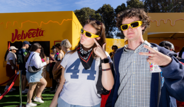 Two people wear yellow sunglasses and pose in front of a yellow Velveeta booth, while other people wait in line nearby on a sunny day.
