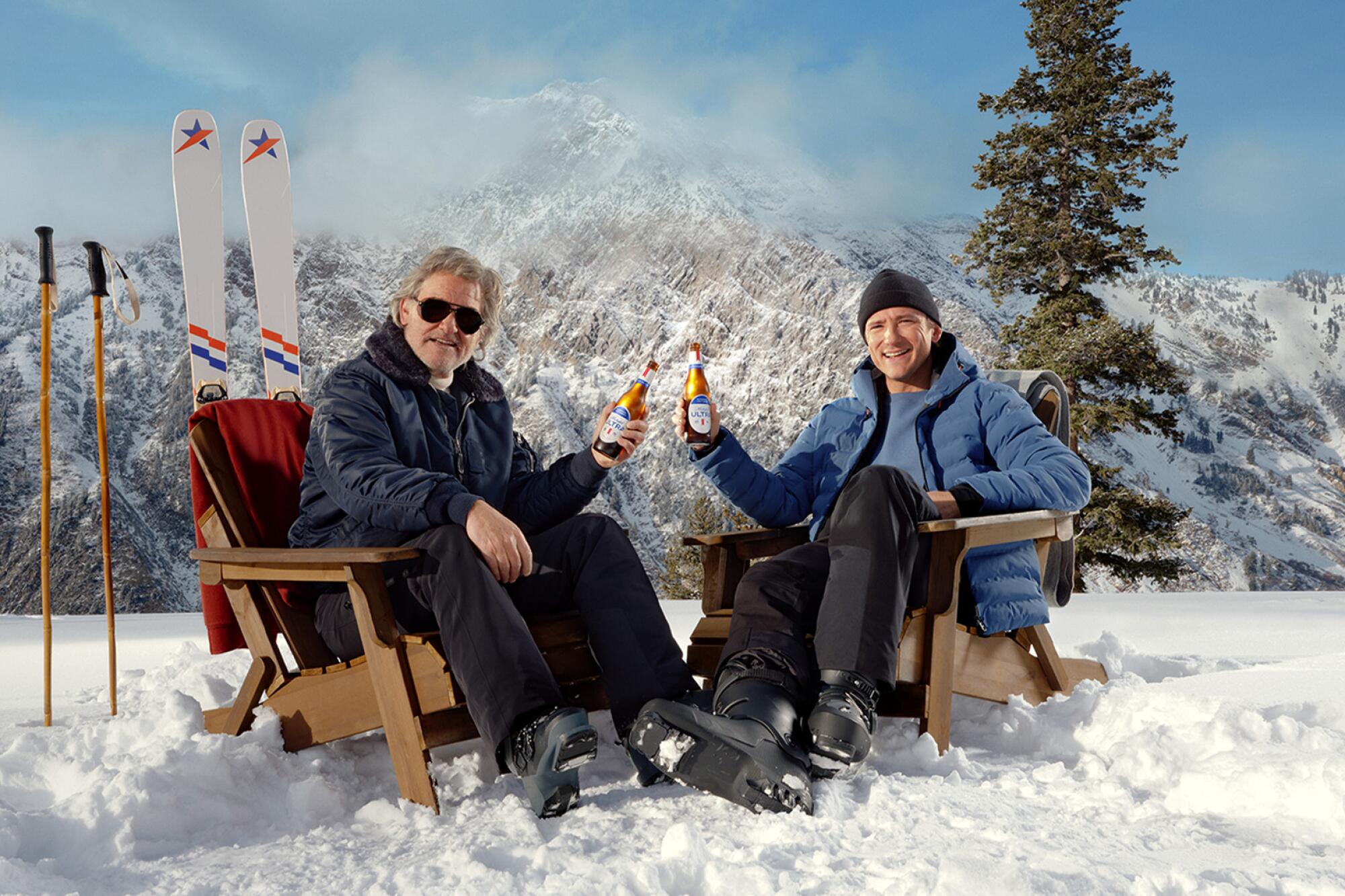 Two men sitting in brown adirondack chairs holding beers with a set of skis and mountains behind them.