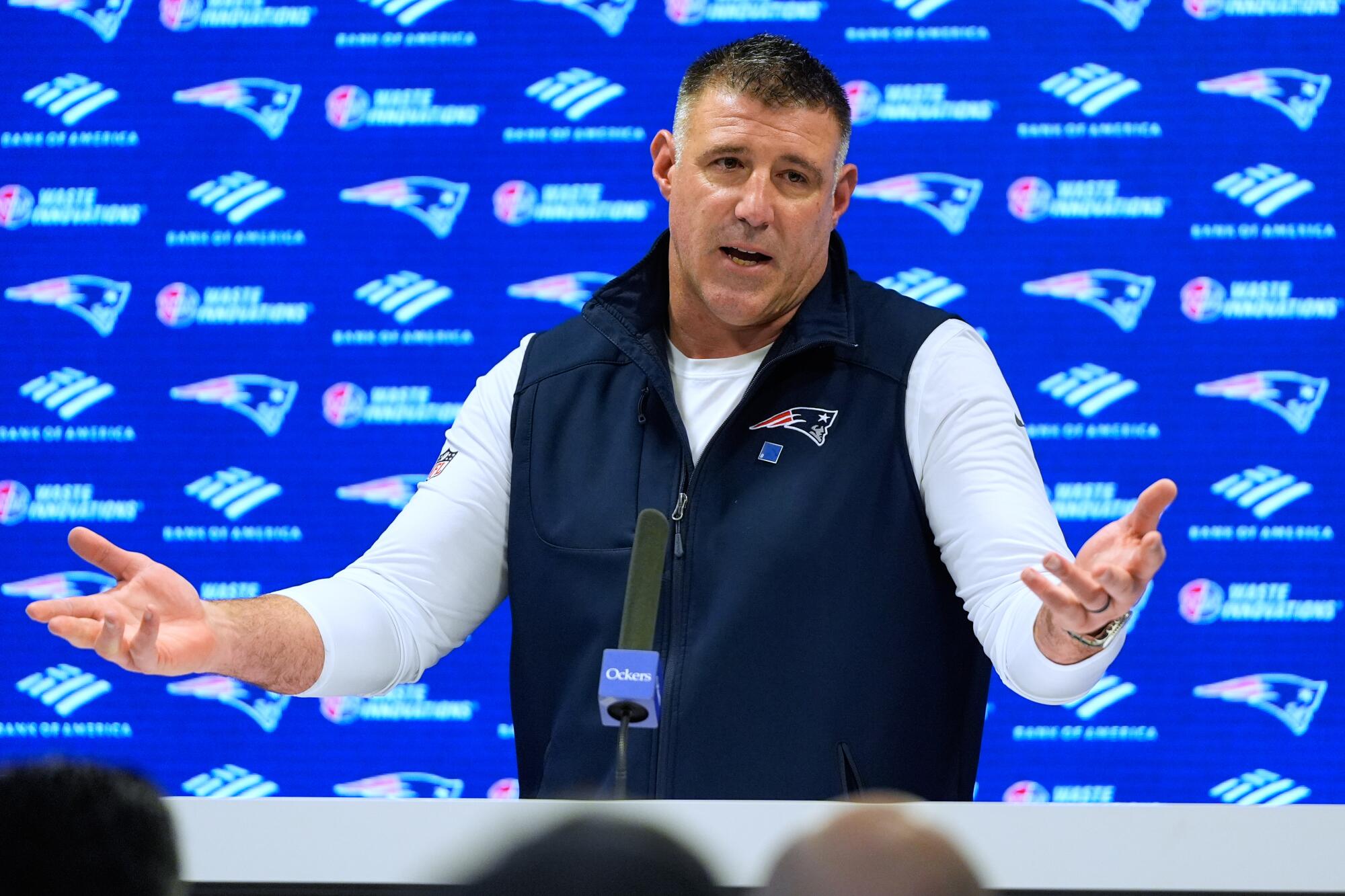 New England Patriots coach Mike Vrabel spreads his arms as he speaks to reporters from a podium.