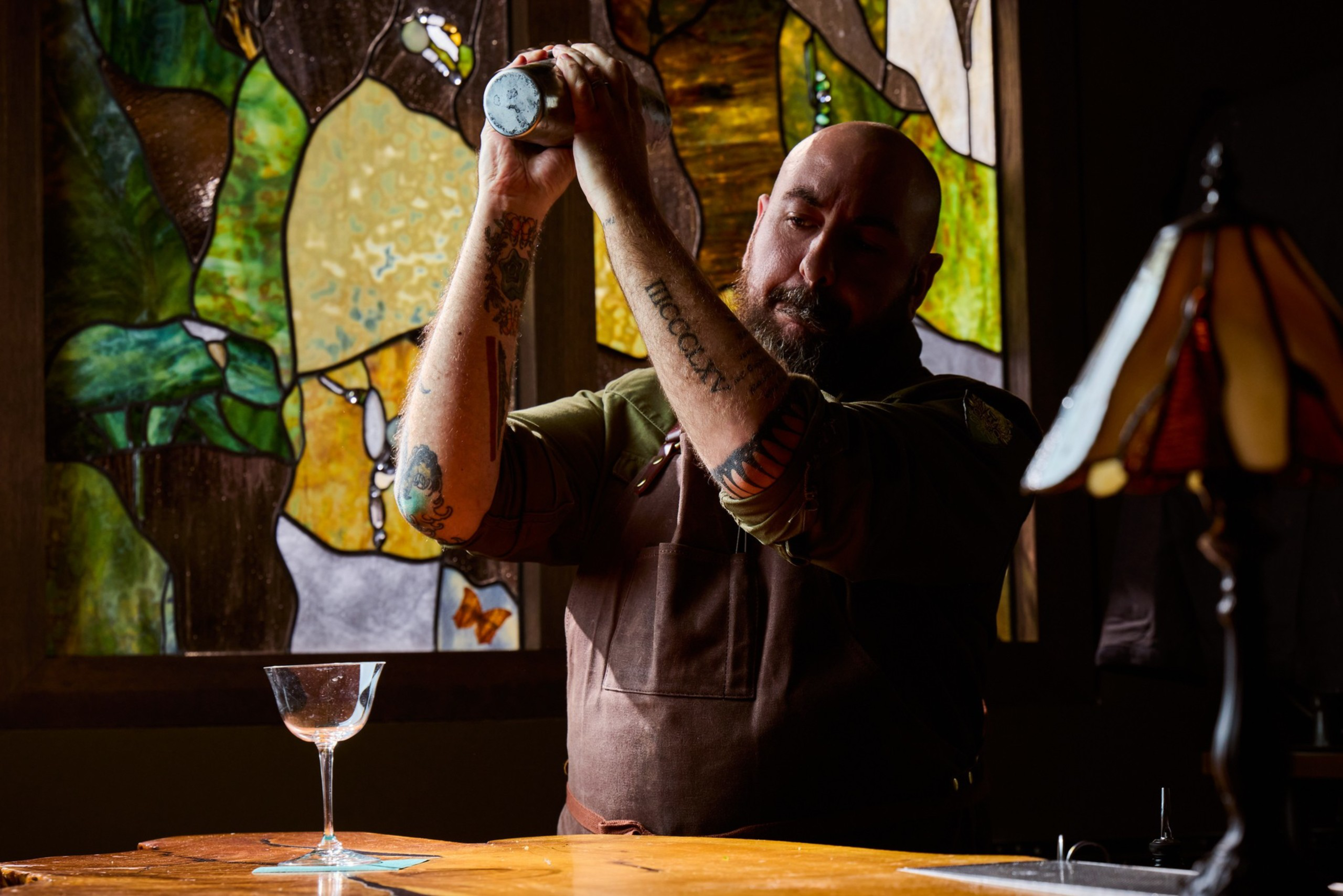 A tattooed man with a bald head and beard shakes a cocktail shaker behind a wooden bar with a stained glass window and an empty glass nearby.