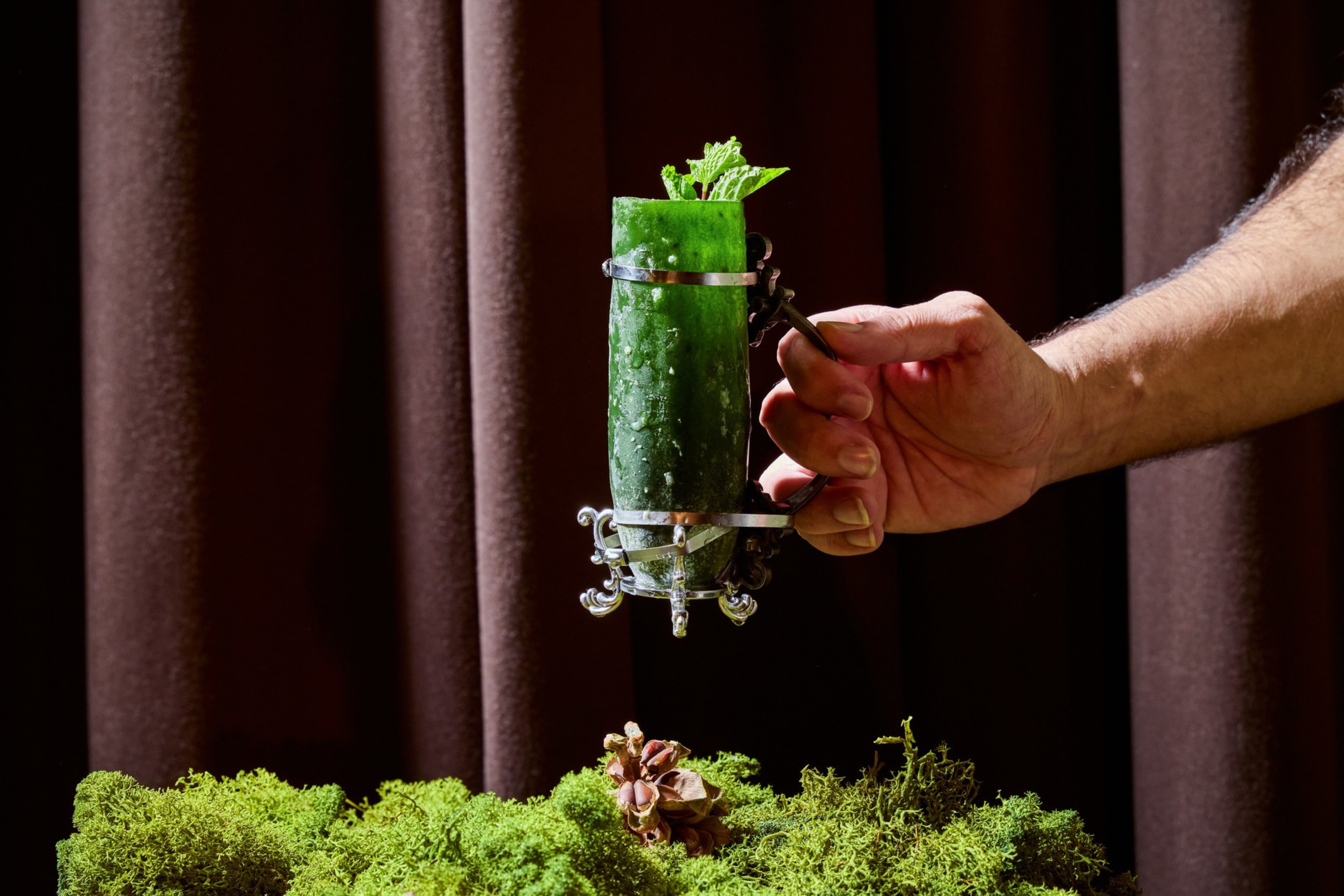 A hand holds an ornate silver cup containing a tall cucumber garnished with mint leaves above a bed of green moss and a pinecone.