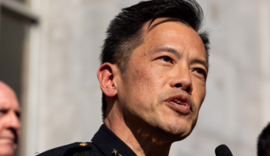 A man in a dark uniform with gold stars on the collar speaks into a microphone, with two blurred people visible in the background.