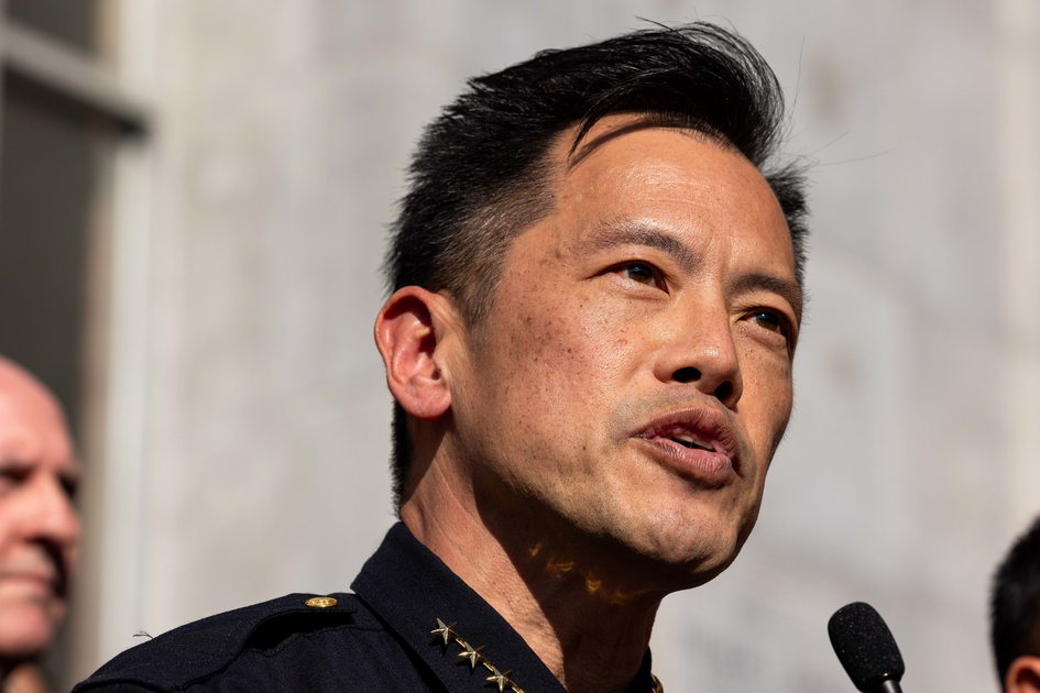 A man in a dark uniform with gold stars on the collar speaks into a microphone, with two blurred people visible in the background.