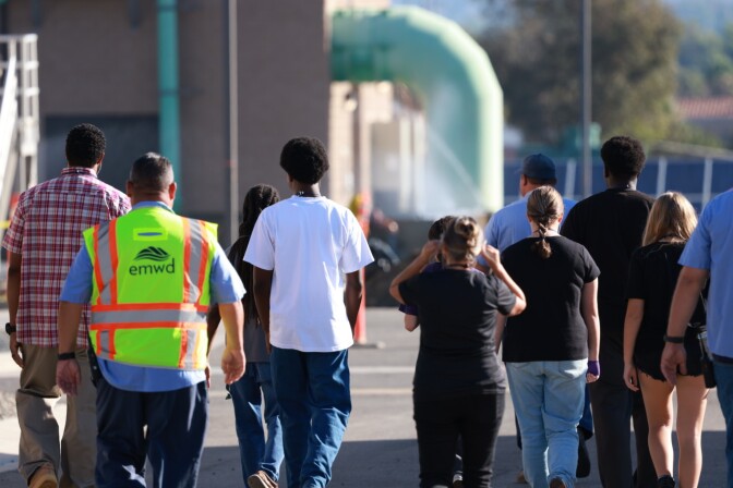 A group of students and an adult wearing a reflective jacket that reads "EMWD" walk away from the camera outside on a sunny day at a water treatment facility.